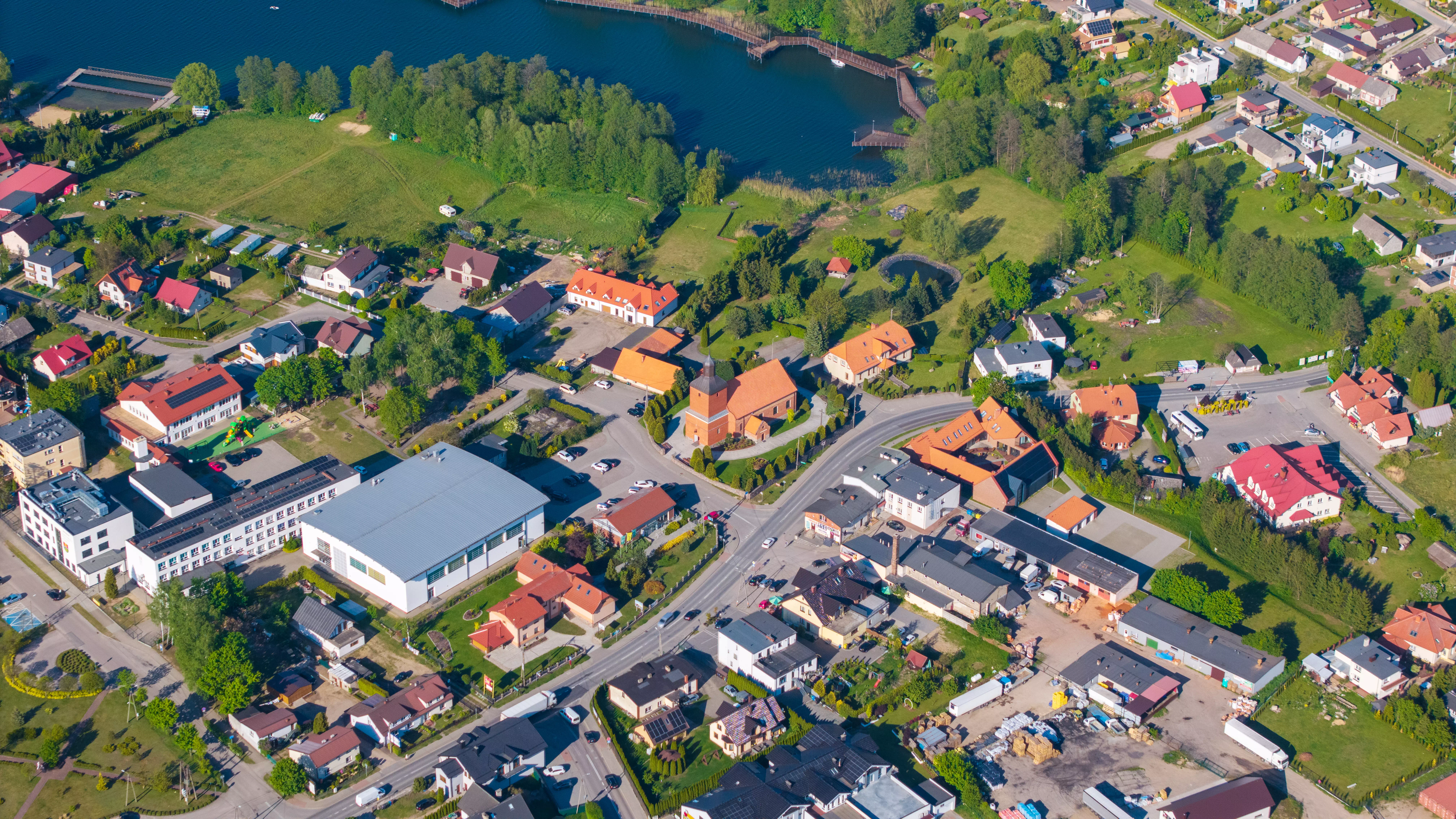 Panorama jeziora otoczonego lasami i polami, z widocznym stadionem i zabudową jednorodzinną