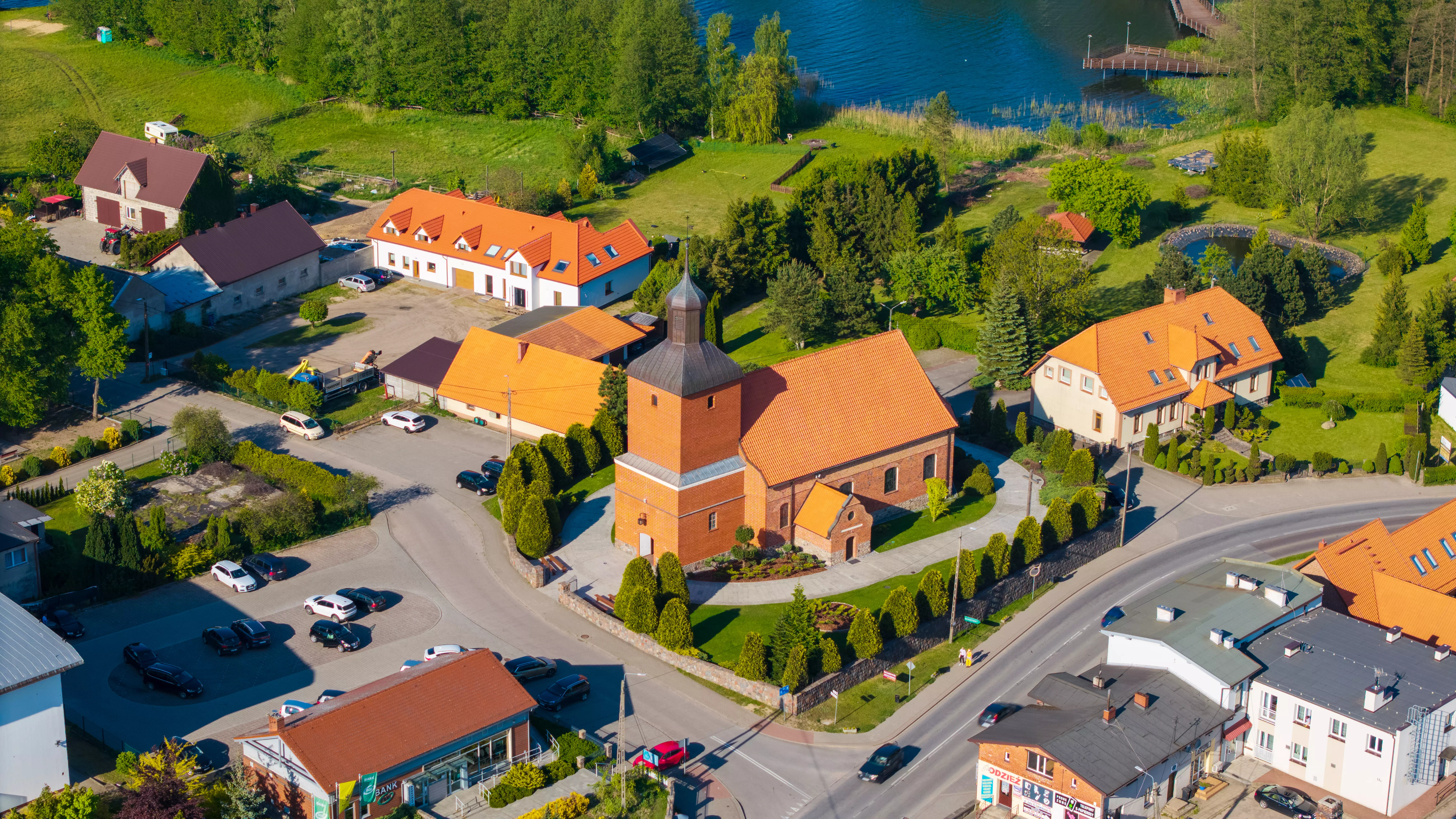 Panorama miejscowości z licznymi domami jednorodzinnymi, polami uprawnymi i jeziorami w tle, widok z lotu ptaka