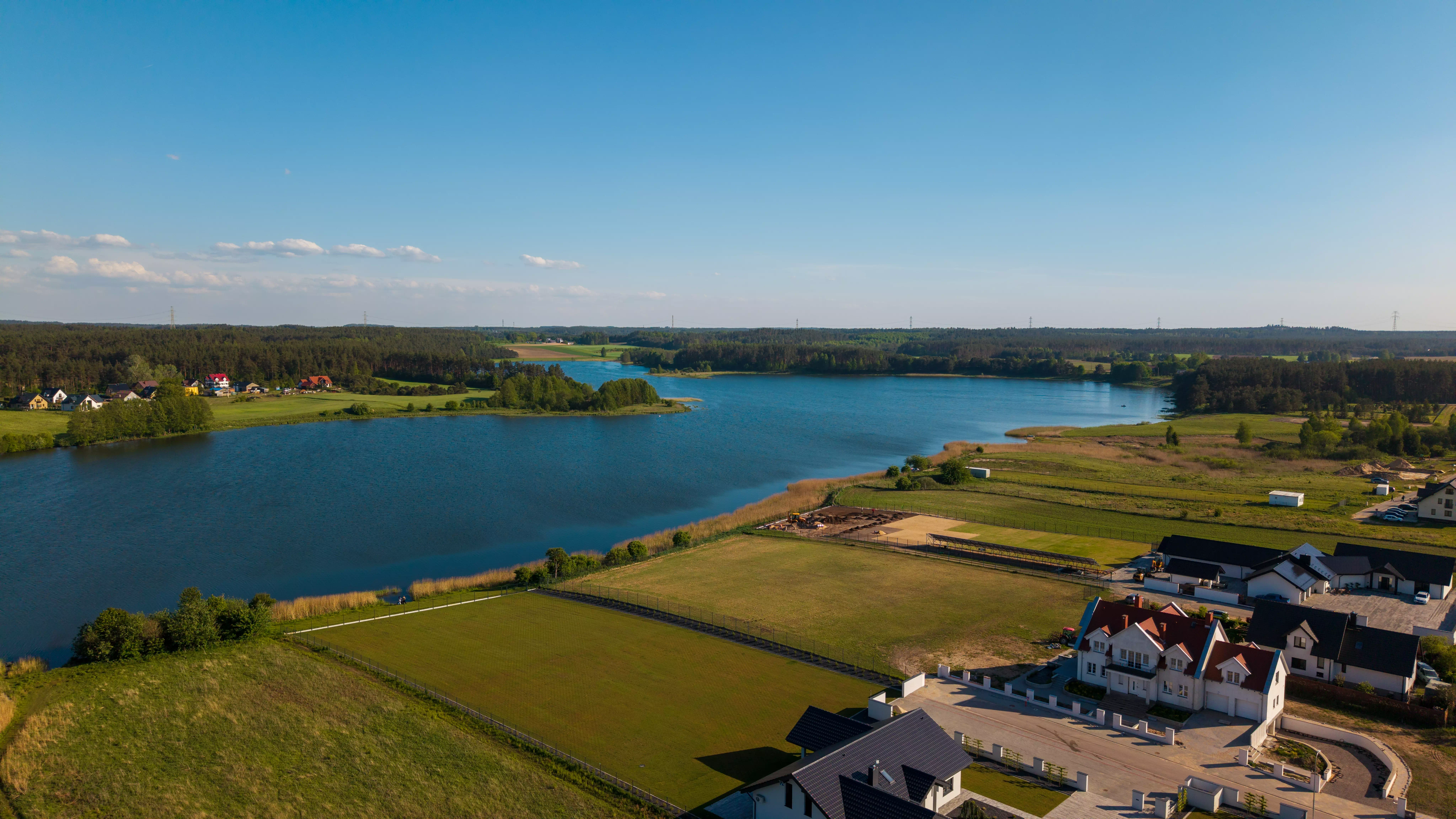 Nowoczesny dom z cegły i białe domy jednorodzinne nad jeziorem, w tle panorama miejscowości i lasy, widok z lotu ptaka
