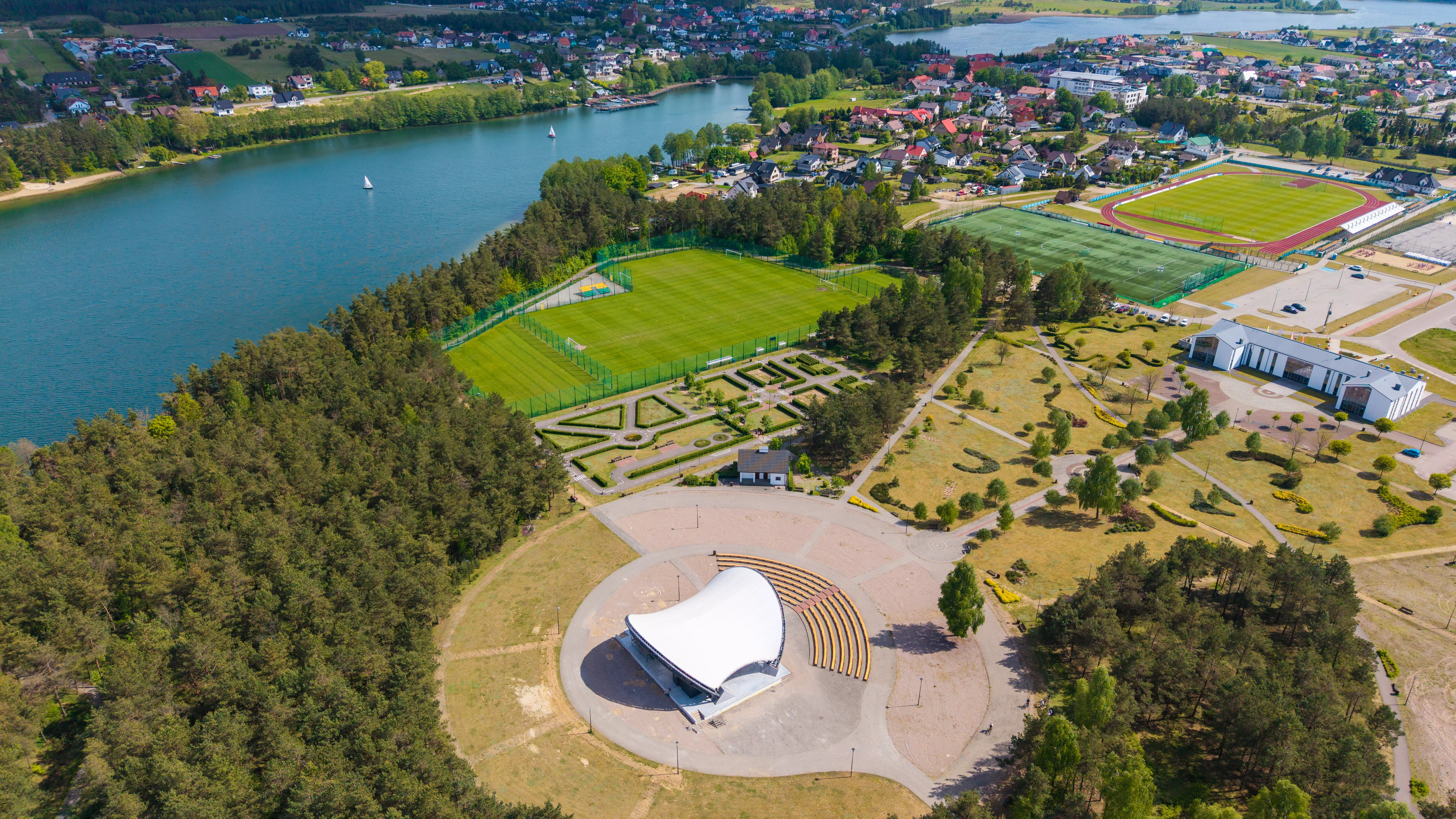 Amfiteatr, park oraz boiska sportowe nad jeziorem w otoczeniu lasów i zabudowy miejskiej, zdjęcie z lotu ptaka