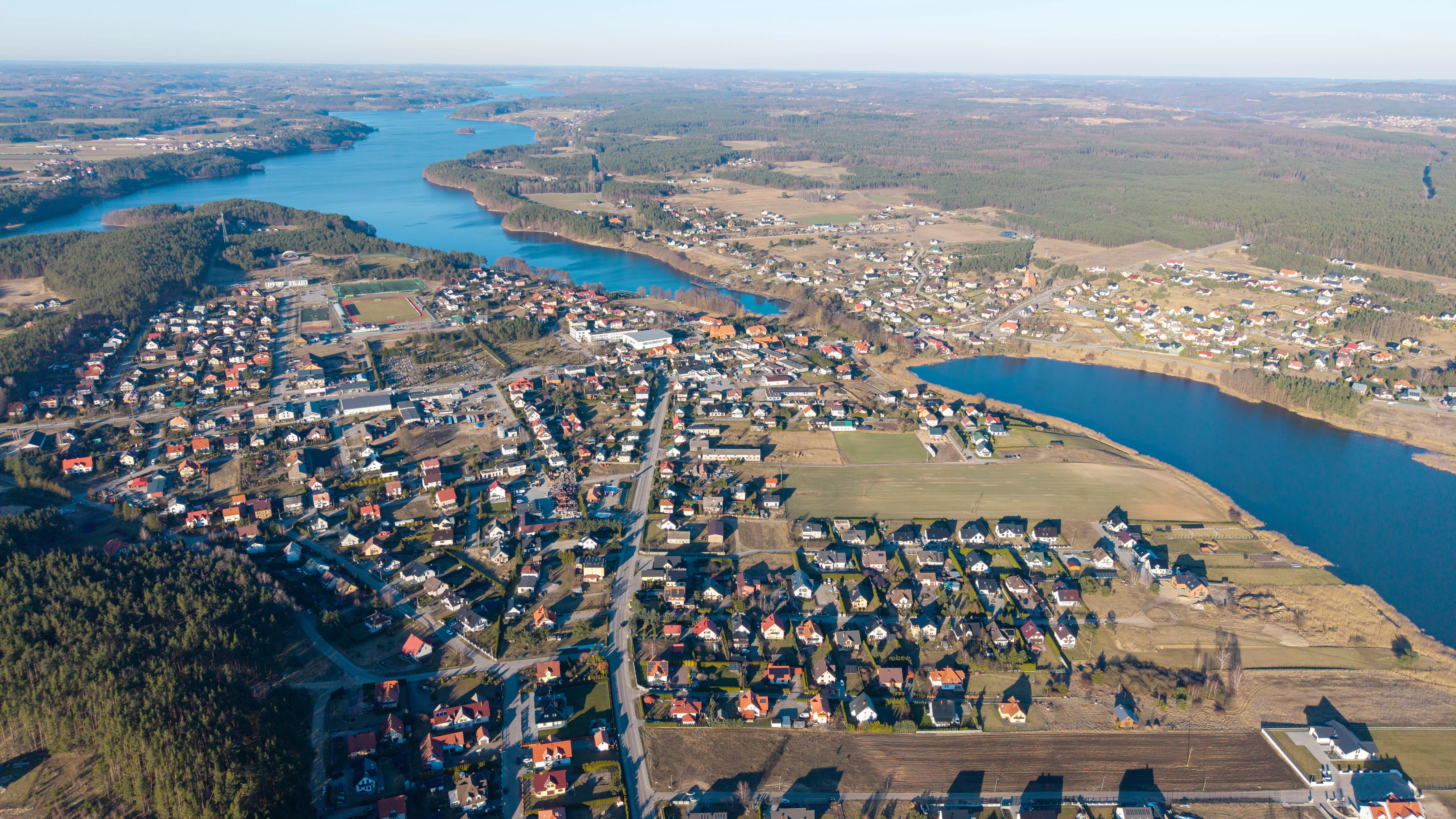 Panorama miasta z widocznym stadionem, zabudową jednorodzinną, jeziorem i otaczającymi lasami, widok z lotu ptaka