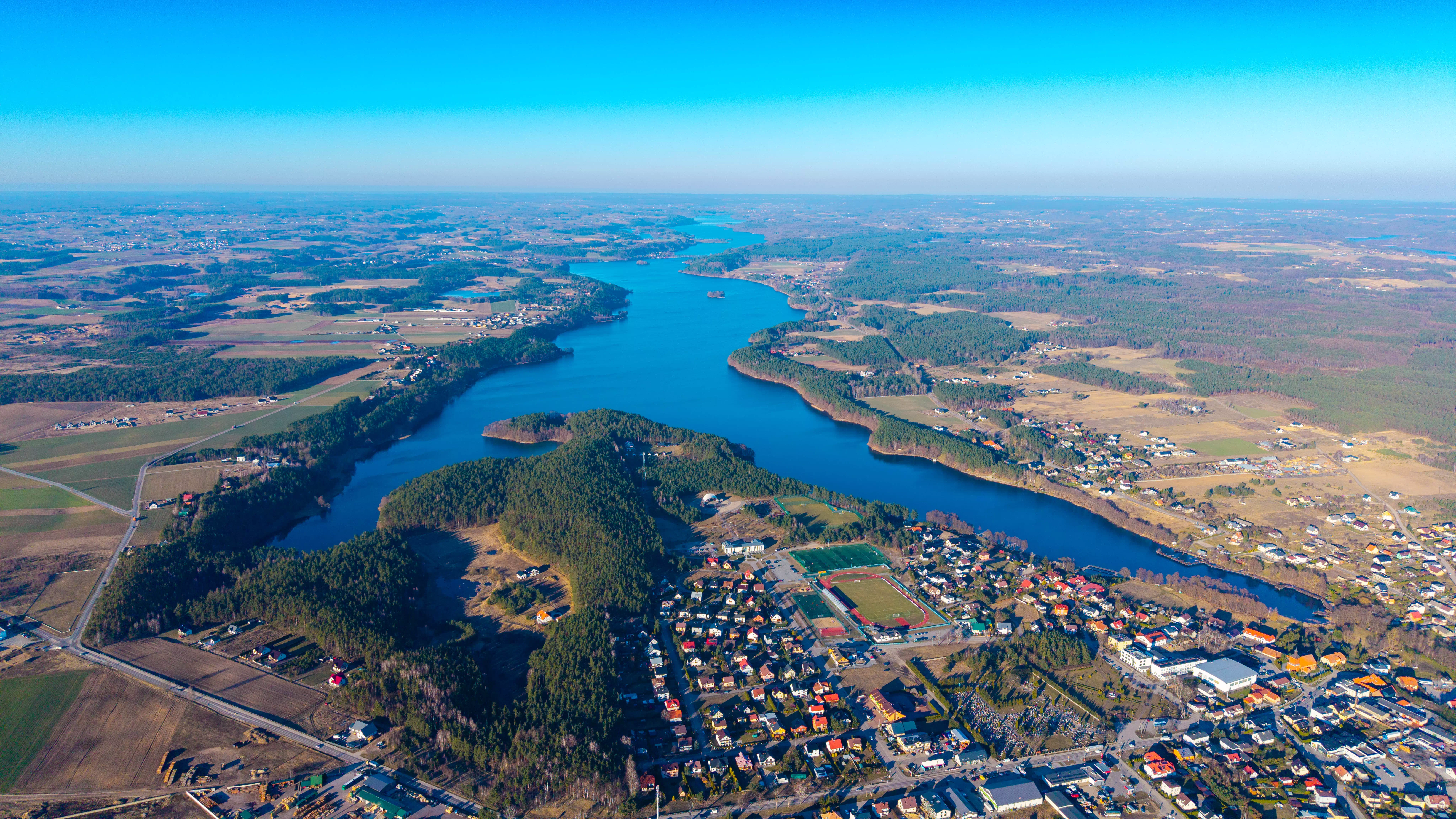 Jezioro otoczone polami uprawnymi, lasami i zabudową wiejską, widok z lotu ptaka