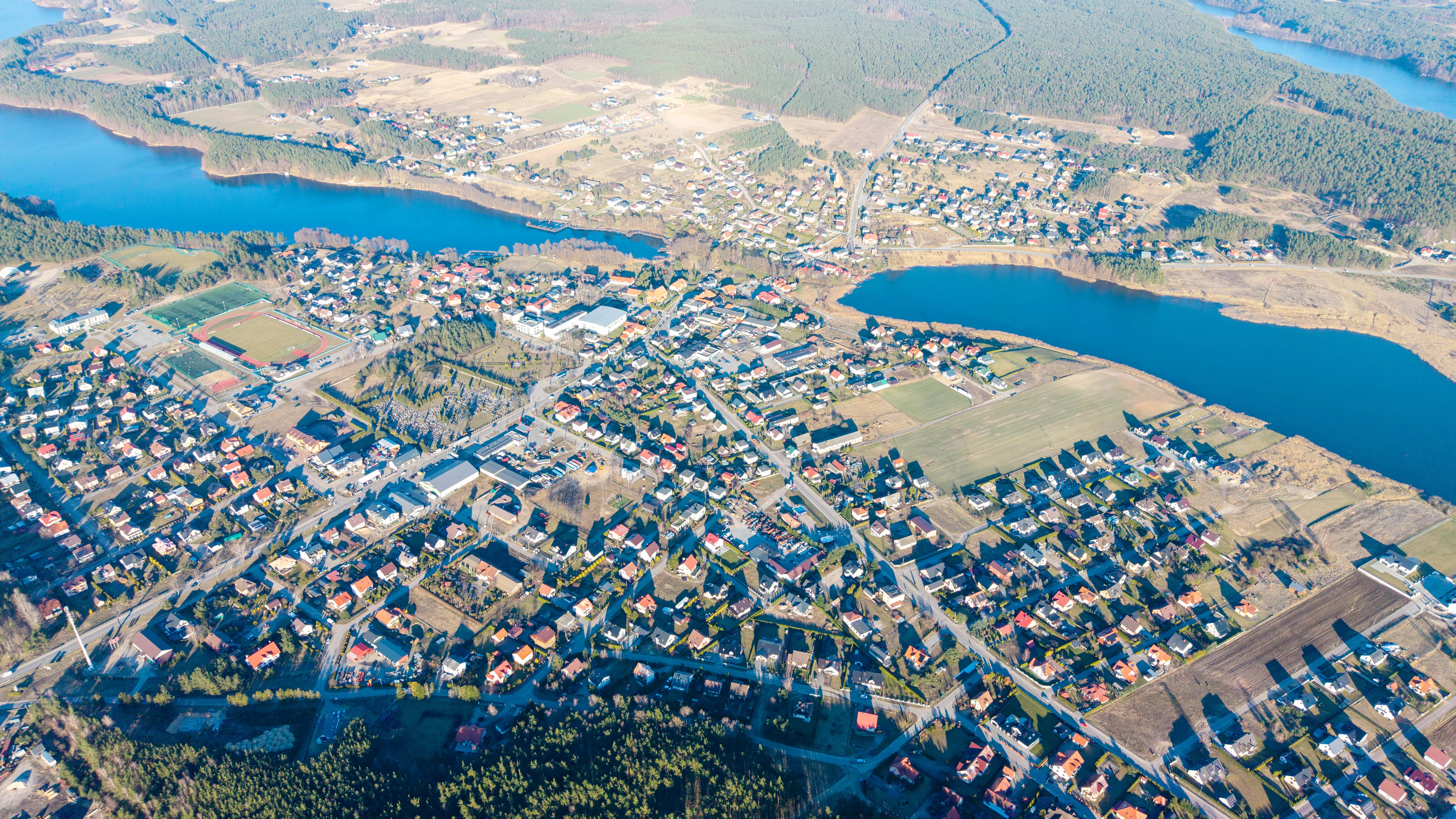 Panorama jeziora wijącego się przez lasy i pola, z fragmentem miasta i stadionem w dolnej części zdjęcia, widok z lotu ptaka