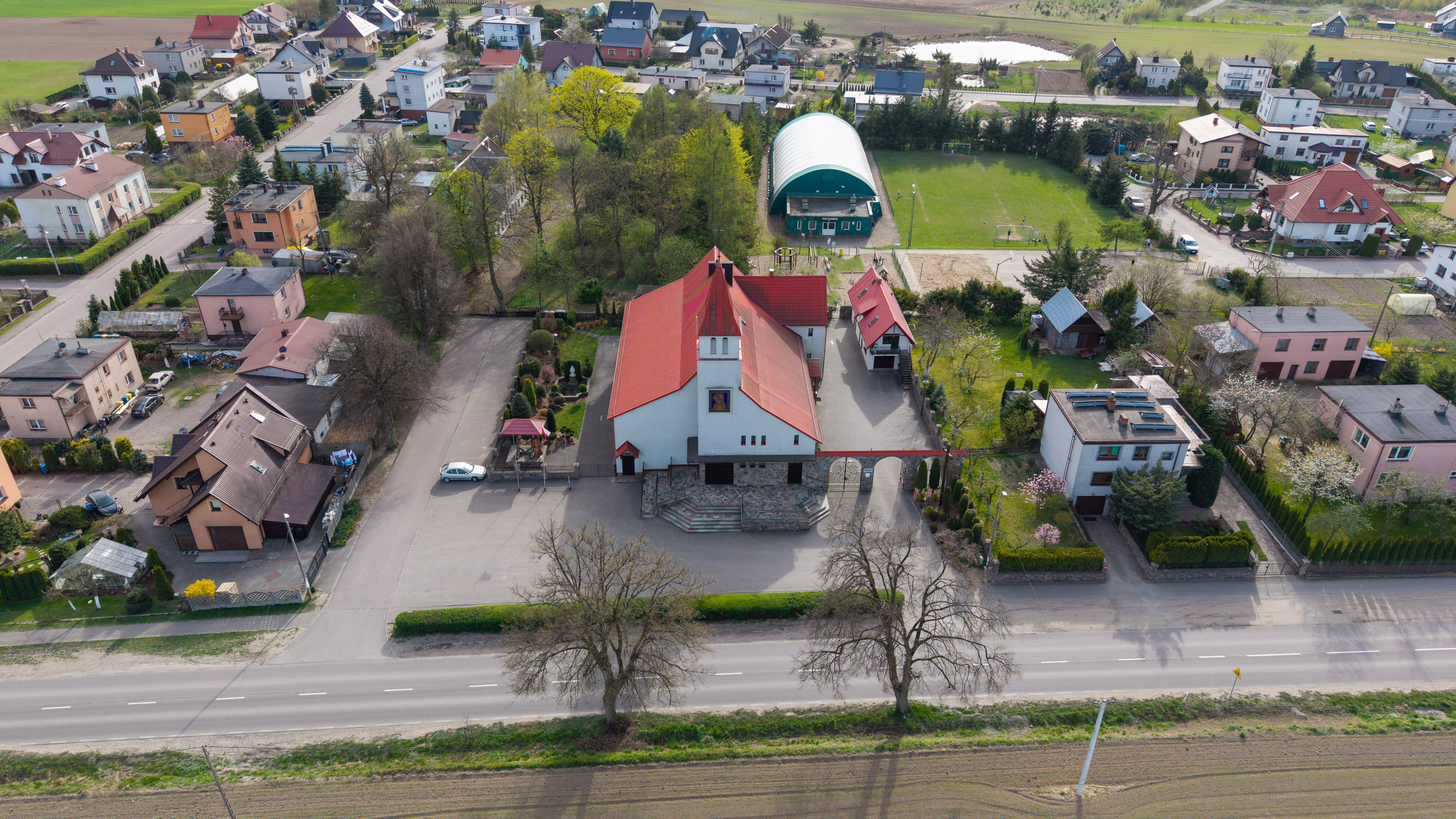 Widok z lotu ptaka na kościół z czerwonym dachem otoczony domami jednorodzinnymi i boiskiem sportowym w małej miejscowości